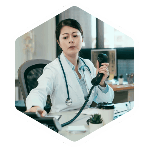 Ärztin sitzt am Schreibtisch, hält ein Telefonhörer ans Ohr und greift gleichzeitig nach einem anderen Telefon.