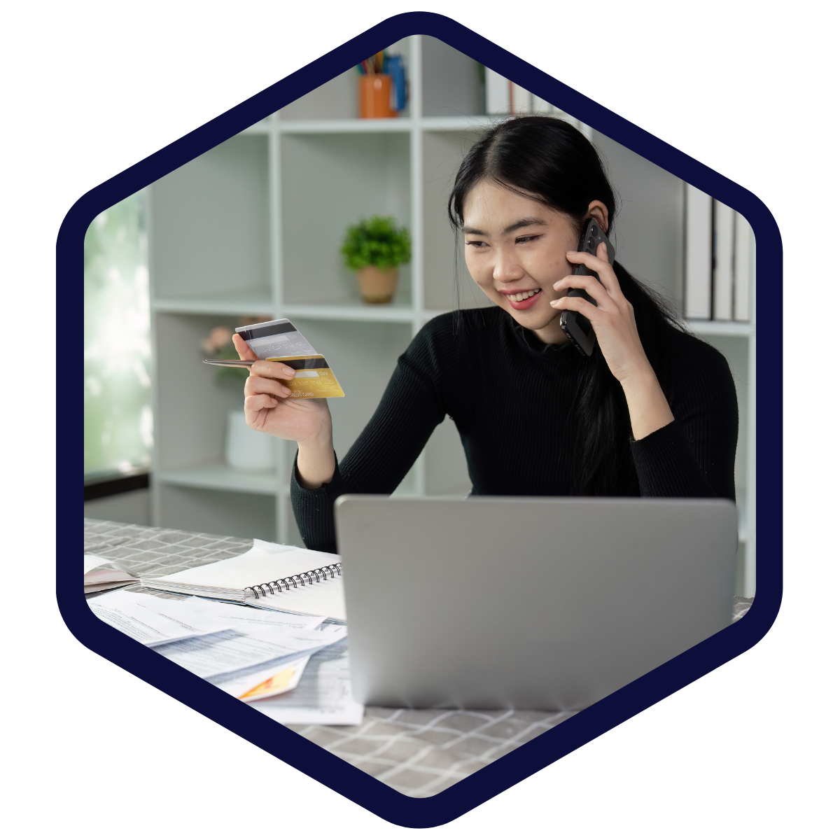 Frau sitzt am Tisch vor einem Laptop, telefoniert und hält zwei Kreditkarten in der Hand; auf dem Tisch liegen Unterlagen.