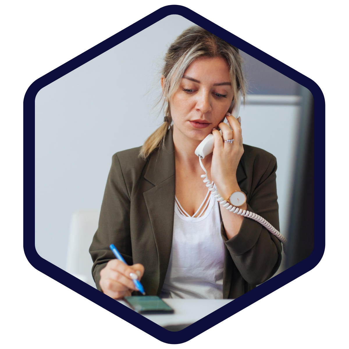 Frau telefoniert mit einem Festnetztelefon und macht gleichzeitig Notizen auf einem Notizblock; sie trägt einen dunkelgrünen Blazer.