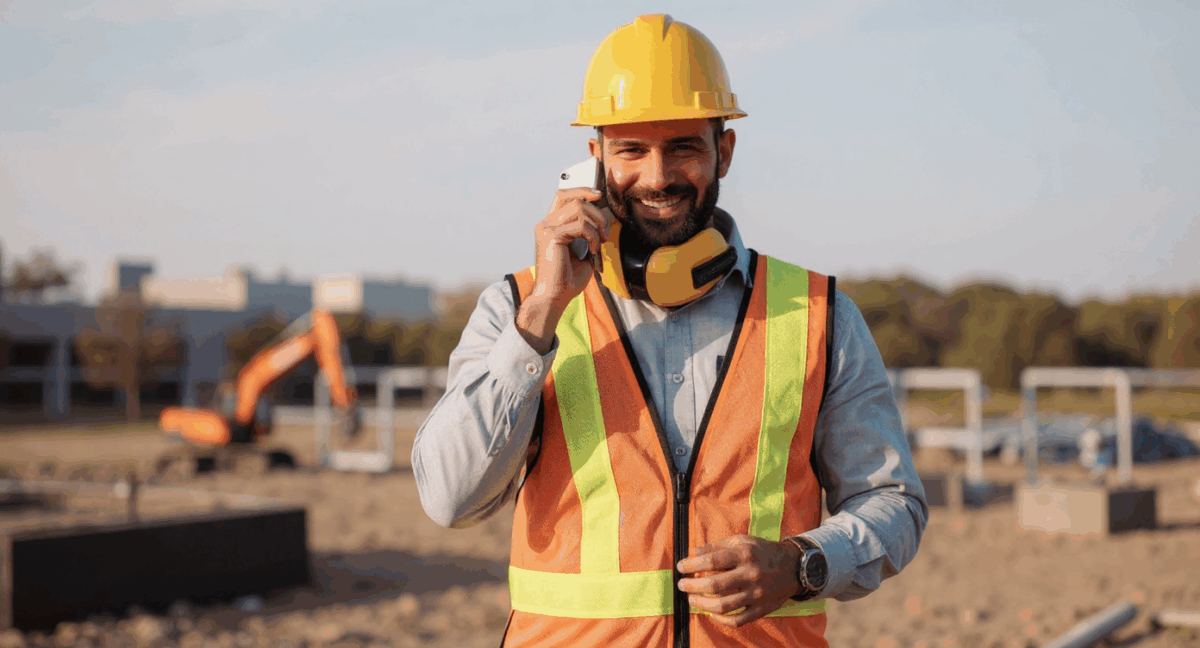 Ein Bauarbeiter mit gelbem Helm, orangefarbener Warnweste und Gehörschutz um den Hals telefoniert lächelnd auf einer Baustelle; im Hintergrund sind Baumaschinen und Gebäudestrukturen zu sehen.